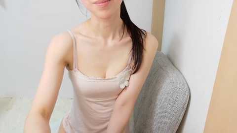 Video of a slender, light-skinned Asian woman with dark hair tied back, wearing a pink spaghetti-strap tank top, sitting on a gray textured chair. She has small breasts and a neutral expression. The background features white walls and a beige floor.