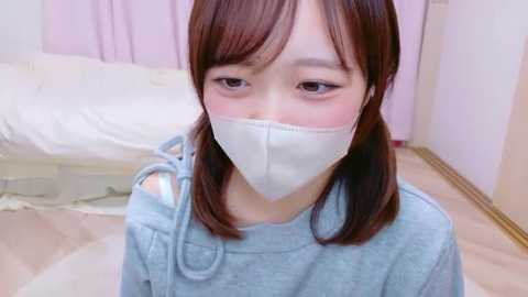 Video of a young Asian woman with straight brown hair, wearing a light blue sweater and white face mask, sitting indoors on a wooden floor with a bed and pastel curtains in the background.