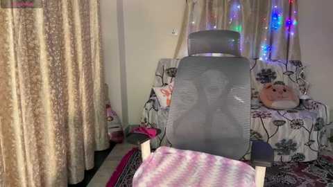 Video of a cozy bedroom with a gray mesh office chair facing a floral-patterned sofa, adorned with stuffed toys and colorful fairy lights. The background features beige curtains and a pink blanket.