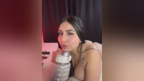 Video of a topless young woman with long dark hair, drinking milk from a glass bottle, sitting on a bed with a black curtain backdrop.