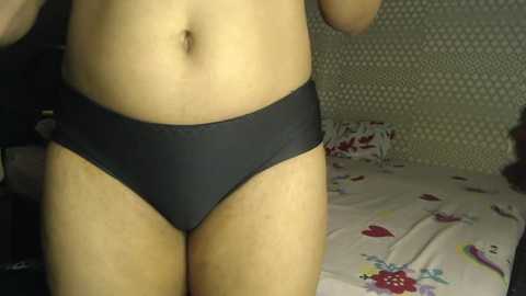 Video of a light-skinned person wearing black panties, standing in a bedroom with patterned wallpaper and a bed with floral bedding.