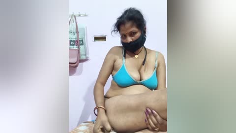 Video of a plus-sized South Asian woman in a blue bra, black mask, and black leggings, sitting on a bed, with a pink handbag hanging on a wall behind her.