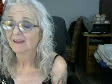 Video of a middle-aged woman with silver hair, wearing glasses, black top, and tattoos, smiling in a cluttered room with a desk, chair, and wicker basket.