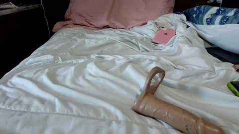 Video of a messy, unmade bed with wrinkled white sheets and a pink pillow, featuring a brown handbag and a pink smartphone.