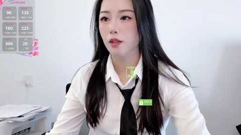 Video of an East Asian woman with long black hair, wearing a white dress shirt, black tie, and green name tag, sitting in a clinical setting with a white wall and medical charts.