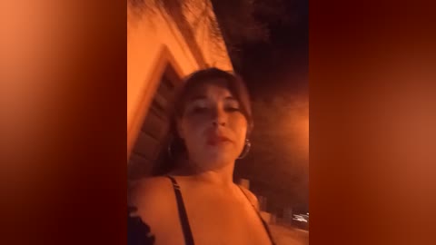 Video of a young woman with medium-length brown hair, wearing a black top, standing in a dimly lit outdoor alley with warm, orange-toned lighting. She appears serene, with closed eyes and a slight smile.