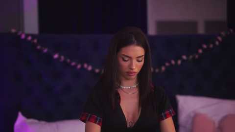 Video of a young woman with long dark hair, wearing a black V-neck top and pearl necklace, seated on a bed with a dark blue tufted headboard, dimly lit with purple ambient lighting.