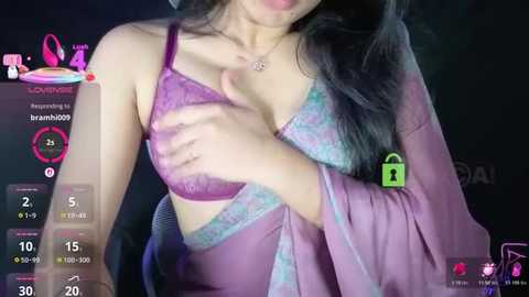 Video of an Asian woman with long black hair, wearing a purple lace bra and sheer robe, partially covering her breasts. The background is dimly lit, with digital overlay elements showing a camera feed and time.