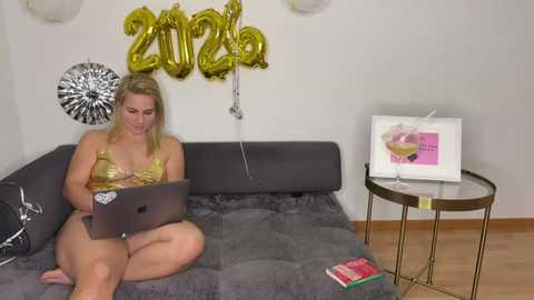Video of a blonde woman in a gold bikini, sitting on a grey couch, typing on a laptop, with a champagne flute, gold \"2020\" balloon, and books on a wooden floor.