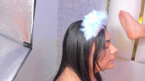 Video of a young woman with long, wet, dark hair wearing a white feather headband, sitting in a bathtub, her feet raised, in a bathroom with white tiled walls and a frosted glass shower door.