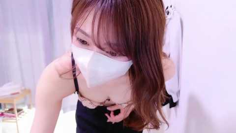 Media: Video of an Asian woman with light skin, wearing a white face mask, black dress, and long brown hair, bending forward in a room with white walls and a white curtain.
