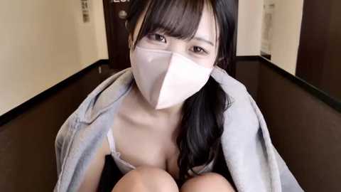 Video of an East Asian woman with long black hair, wearing a light grey denim jacket over a white bra, sitting on the floor in a narrow hallway with wooden floors and dark walls.