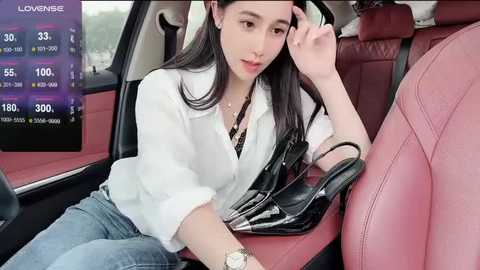 Video of an East Asian woman with long black hair, wearing a white blouse and blue jeans, seated in a pink leather car seat. She holds a black leather handbag.