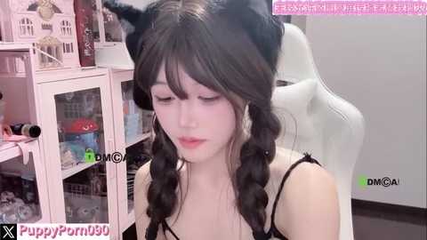 Video of a young Asian woman with long, black hair styled in twin braids, wearing black cat ears and a black lace bra. She sits in a white gaming chair in a pink-themed room with shelves and toys.