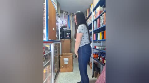 Media: Video of a woman with long black hair, wearing a gray shirt and blue jeans, standing in a cluttered, brightly-lit convenience store, looking at a display case filled with snacks and drinks.