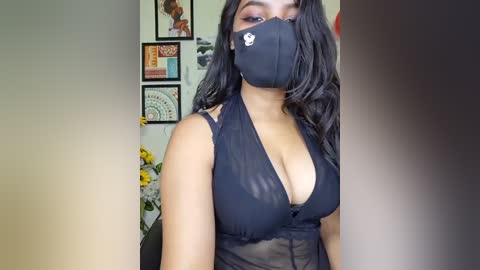 Video of a young woman with medium brown skin, wearing a black face mask and sheer black lingerie. She stands in front of a wall adorned with framed art and flowers, creating a cozy, intimate setting.