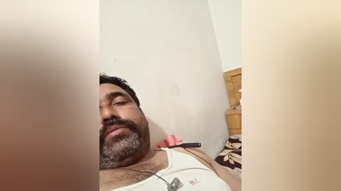 Video of a middle-aged South Asian man with a beard and dark hair, lying on a bed with a white tank top, eyes closed, in a dimly lit room.