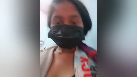 Media: Video of a woman with short dark hair, wearing a black face mask, gray hoodie, and a colorful scarf with red text, in a room with light blue walls.