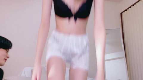 Video of a slender, fair-skinned woman wearing a black bra and white lace panties, standing in a bedroom with a bed and beige curtains in the background.