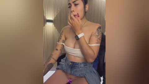Video of a Latina woman with tattoos, wearing a strapless top and denim skirt, sitting on a bed, touching her lips suggestively.