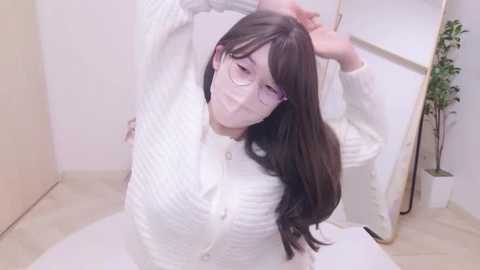 Media: Video of a young Asian woman with long black hair, glasses, and a white sweater, stretching in a modern, minimalist room with a white wall and plant.