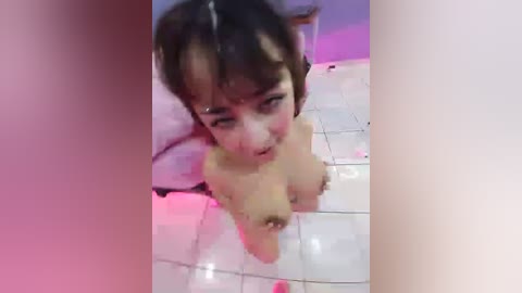 Video of a young Asian woman with short, dark hair, lying naked on a white tiled floor with a pink background, her breasts exposed and face slightly flushed.
