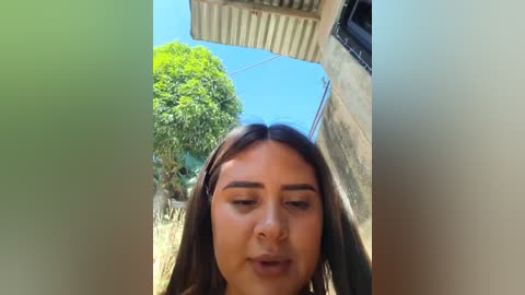 A video of a young woman with long dark hair, closed eyes, and a slight smile, standing in a sunlit alleyway with a lush tree and rustic buildings in the background.