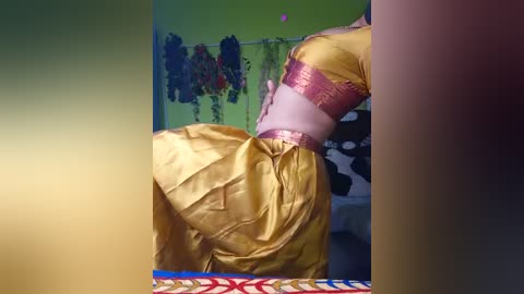 Video of a person in a golden, satin sari with a purple border, standing against a green wall adorned with colorful garlands. The sari's texture is smooth and glossy, highlighting the elegant folds.