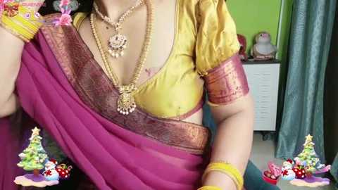 A video of a South Asian woman wearing a yellow blouse, purple sari, gold necklace, and bracelets, set in a colorful, festive room with green walls, a blue curtain, and a pink statue.