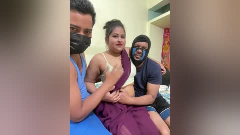 Video of three people in a room: a woman in a beige bra and purple skirt, a man in a black mask and blue shirt, and another man in a blue shirt, all smiling.