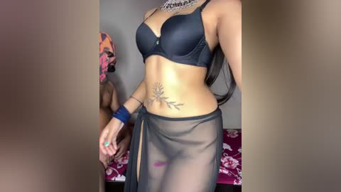 A video of a slender woman in black lingerie, including a bra and sheer wrap skirt, standing in a bedroom with a floral bedspread. She has a tattoo on her abdomen.
