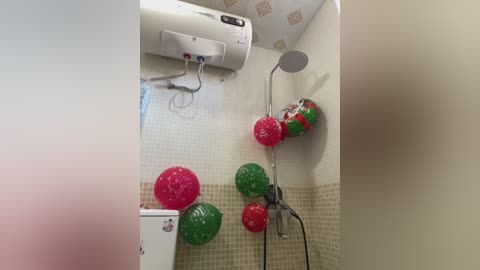 Video of a small bathroom with white tiled walls, a white toilet, and a white wall-mounted heater. The walls are decorated with festive red and green balloons and a decorative showerhead, creating a holiday-themed ambiance.