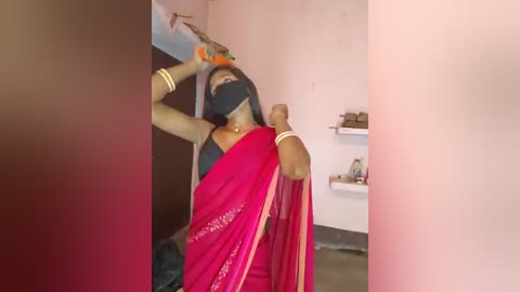 Media: Video of a South Asian woman in a red saree, black mask, and gold bangles, dancing in a brightly lit room with white walls and shelves.