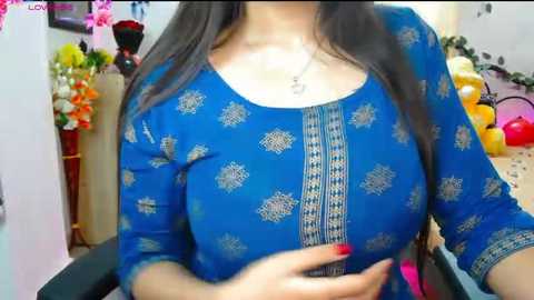 Video of a woman in a blue, patterned kurta with intricate embroidery, wearing a pendant necklace. Background includes flowers, balloons, and a table.