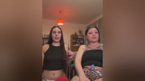 Video of two women, one slim with long dark hair, wearing a black crop top, the other heavyset with short dark hair, wearing a leopard-print top, sitting on a couch, in a cozy room with shelves and a red lamp.