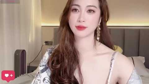 A video of a young East Asian woman with long, wavy brown hair, fair skin, and full red lips. She wears a patterned off-shoulder top and dangling earrings. The background features a modern, minimalist bedroom with a grey headboard.