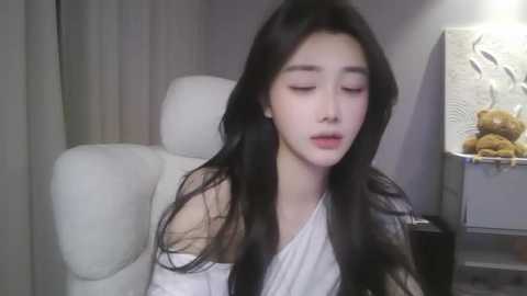 Video of a young Asian woman with long, straight black hair and fair skin, wearing a white off-shoulder top, sitting on a white leather chair in a modern, minimalist room with a decorative wall panel and a vase of stuffed animals.