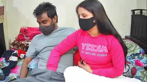 Video of a man and woman wearing masks on a bed with colorful blankets, discussing in a simple room.