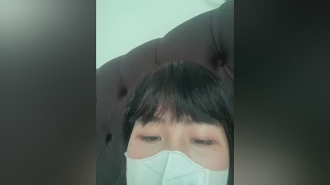 A close-up video of an Asian person with short black hair, wearing a white surgical mask, lying on a dark-colored, tufted leather sofa. The background is out of focus, with a soft, blurred light source.