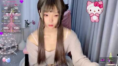 Video of a young Asian woman with long brown hair, wearing a white off-shoulder top, sitting in a pink chair. Background includes a white curtain and a Hello Kitty character.