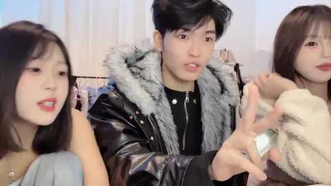 Video of three young Asian people indoors, wearing casual clothes. Man in black leather jacket and fur collar, woman with red lips on left, woman with straight brown hair on right.