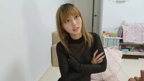 A young Asian woman with straight, shoulder-length light brown hair and fair skin sits on a beige chair in a minimalist bedroom, wearing a sheer black top. The background features a white wall, a pink quilted bed, and a cluttered desk with books and a lamp.