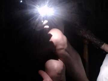 A video captures a dimly lit room with two people, one bald and another with long hair, both illuminated by a bright flashlight. The room's background is dark and indistinct.