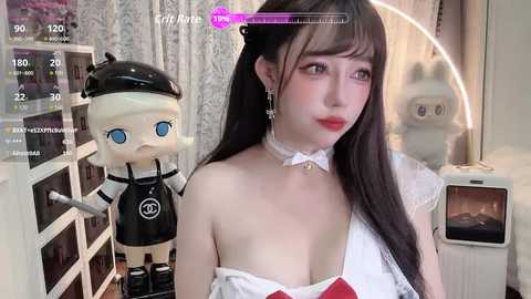 A video of an East Asian woman with long black hair, wearing a white off-shoulder dress, a choker, and earrings, standing next to a plush toy in a room with white walls and a television.