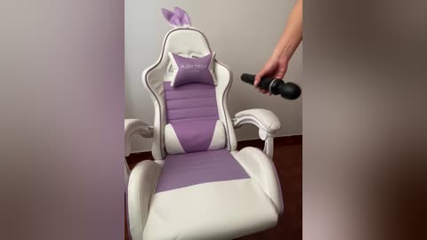 Video of a white gaming chair with purple accents and \"Naughty\" pillow, being massaged by a hand holding a black massage wand, in a minimalist room with beige walls and wooden floor.