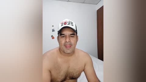 A video of a shirtless, slightly overweight Asian man with a medium build, wearing a white baseball cap with red and black writing, in a simple, white-walled room.