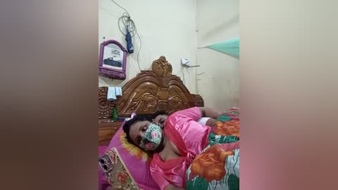 Video of an Indian woman with dark skin and short hair, wearing a pink sari and face mask, lying in a small, dimly lit room with a wooden headboard, colorful bedspread, and a framed picture on the wall.