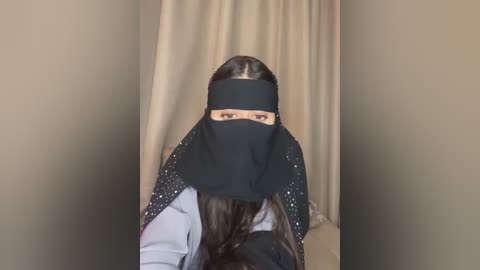 Video of a woman in a black hijab and long brown hair, standing in a beige room with neutral-toned curtains, wearing a light gray shirt.