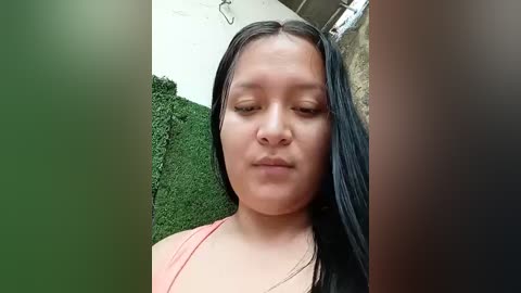 Video of a Latina woman with long black hair, light skin, and a neutral expression, wearing a pink top, standing in front of a green hedge and concrete wall.