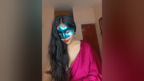 A video of a young woman with long black hair, wearing a vibrant blue mask and a pink sari, standing in a dimly lit room with beige walls and a wooden door.